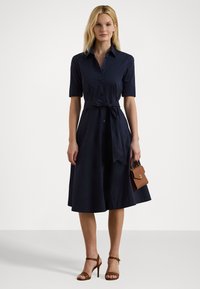 Navy shirt dress with a fitted bodice, short sleeves, and a tie at the waist; paired with brown heeled sandals and a small handbag.