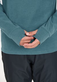 Turkoosi pitkähihaisen paidan, jossa on peukaloaukot, pehmeä rakenne ja rento mitoitus. Kädet ovat ristissä edessä, mikä näyttää hihansuiden suunnittelun.