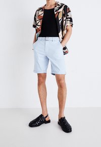 Chemise à manches courtes à motifs avec des couleurs noire, beige et rouille, associée à un short bleu clair avec des revers et des sandales noires texturées.