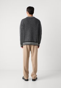 Svart strikkegenser med hvitt geometrisk mønster, kontrasterende stripete kant nederst, båret med beige bukser og svarte sko.
