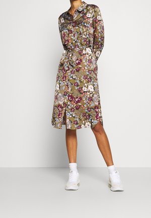 Blumenhemdkleid in Olivgrün mit pinken und weißen Blumen, ausgestattet mit einem taillierten Gürtel, langen Ärmeln und einem knielangen Saum. Weiße Sneakers.