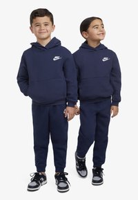 Hoodies da Nike em azul-marinho com logo branco, combinados com calças de treino iguais, usados por duas crianças. Tecido suave, design casual, punhos elásticos.