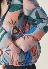 Pufjakke med blomstermønster og bjørneprint i pastelfarver. Lavet af skinnende stof, med justerbar kant og ribbede manchetter.