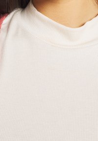 Krémový žebrovaný rolák s vysokým výstřihem, jemnou texturou a subtilním leskem, zaměřující se na kontury a vzhled látky.