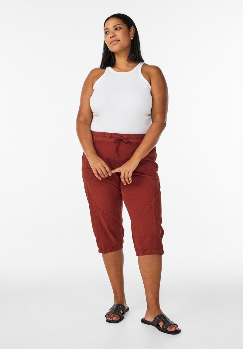 Žena stojaca so spojenými rukami, oblečená v bielom tielku bez rukávov, hnedočervených skrátených nohaviciach a čiernych šľapkách na zapínanie.