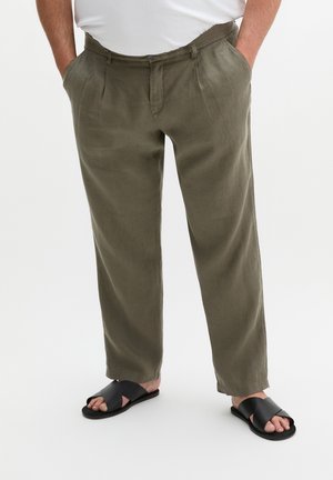 Mann trägt olivgrüne Hose mit den Händen in den Taschen und schwarzen Slide-Sandalen vor weißem Hintergrund.