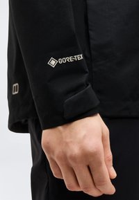Main reposant à côté d'une manche de veste noire portant le logo GORE-TEX brodé sur la zone de l'avant-bras.