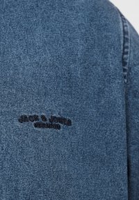 Κοντινό πλάνο μπλε υφάσματος τζιν με μαύρο κεντημένο κείμενο "JACK & JONES PREMIUM" στην περιοχή του άνω μανικιού ενός ενδύματος.