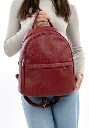 Roter Leder-Rucksack mit strukturiertem Finish, runder Form, vorderer Reißverschlusstasche und doppelten Schultergurten. Von einer Person gehalten.