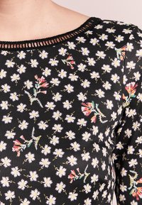 Haut floral noir avec un col large et simple, orné d'une bordure noire, décoré de petites marguerites blanches et d'accents floraux colorés.