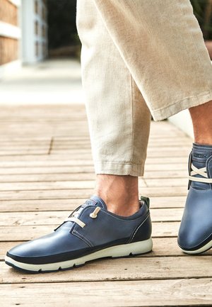 Muž nosící béžové kalhoty a tmavě modré pohodlné tenisky stojí venku na dřevěné podlaze.