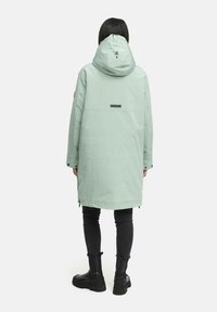 Langer mintgrüner Mantel mit Kapuze, Rückenlabel, Reißverschlusstaschen an den Seiten und einem lockeren Schnitt. Kombiniert mit schwarzen Hosen und klobigen schwarzen Stiefeln.