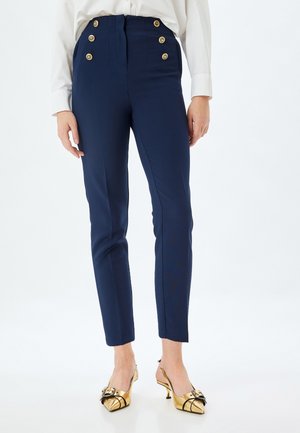 Pantaloni sartoriali blu navy con vita alta, caratterizzati da sei bottoni dorati e un design a gamba affusolata. Tessuto dalla consistenza liscia, abbinato a scarpe con tacco dorate.