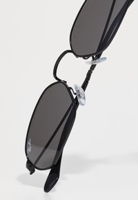 Crne pravokutne sunčane naočale Ray-Ban s tamnim lećama i prozirnim nosnim jastučićima na bijeloj pozadini.