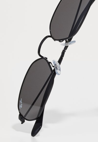 Черни правоъгълни слънчеви очила Ray-Ban с тъмни лещи и прозрачни носни падове на бял фон.