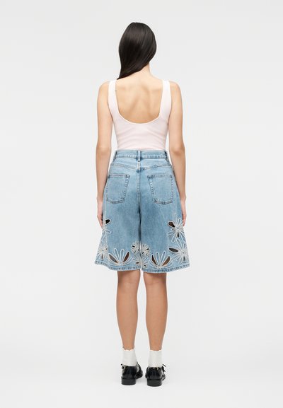 Γυναίκα με μακριά σκούρα μαλλιά, που φορά λευκό αμάνικο τοπ και μπλε τζιν σορτς μέχρι το γόνατο με κεντήματα λουλουδιών με κοψίματα, μαύρα παπούτσια και λευκές κάλτσες.