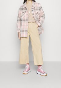Karierte Jacke in Pink, Grau und Weiß, beige weit geschnittene Hose und pinke Sneakers mit weißen Akzenten und dicken Sohlen. Lange Ärmel, zwei Taschen.