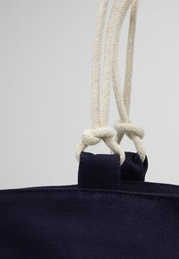 Gros plan sur des poignées en corde torsadée crème nouées à travers des boucles en tissu bleu marine foncé sur un sac en toile, sur un fond gris clair.