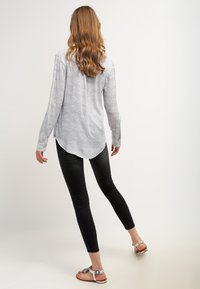 Bluzka z długim rękawem w jasnoszarym kolorze z czarnym nakrapianym wzorem, dopasowany tył, zaokrąglony dół, noszona z czarnymi obcisłymi dżinsami i metalicznymi sandałami.