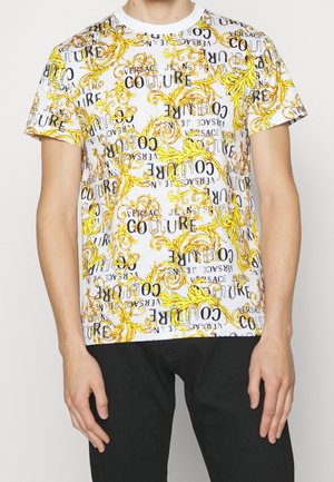 Chemise blanche à manches courtes avec des motifs baroques jaunes et le texte noir répété "VERSACE JEANS COUTURE" portée par une personne en pantalon noir.