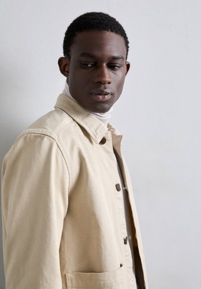 Chaqueta beige clara con diseño de cuello, cierre frontal de botones, tela texturizada y un solo bolsillo en el pecho. Llevada sobre un cuello de tortuga crema.