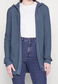 Gebreid vest in marineblauw, met een capuchon, open voorkant en lange mouwen met geribbelde boorden. Draagt over een wit shirt en een spijkerbroek.