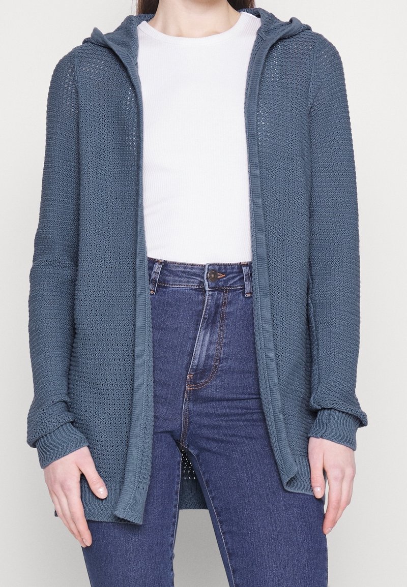 Gebreid vest in marineblauw, met een capuchon, open voorkant en lange mouwen met geribbelde boorden. Draagt over een wit shirt en een spijkerbroek.