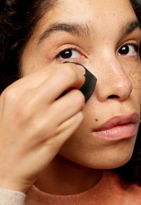 Esponja de maquillaje, negra, aplicando producto debajo del ojo. Primer plano de la piel con pecas y un color de labios suave, con una textura lisa y detalles sutiles.