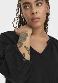 Black top with a V-neckline and long, gathered sleeves. Features a textured fabric and a button detail on the cuff. Subtle tattoo visible on arm.