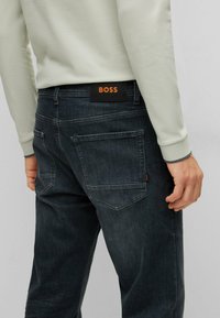 Bakifrån av en person som bär urtvättade svarta jeans med BOSS-logopatch och en ljusbeige långärmad skjorta, stående mot en enfärgad bakgrund.