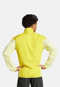 Gelbe leichte Jacke mit hohem Kragen, kontrastierenden hellgelben Ärmeln und glatter Textur. Hat ein tailliertes Design ohne sichtbare Beschläge.