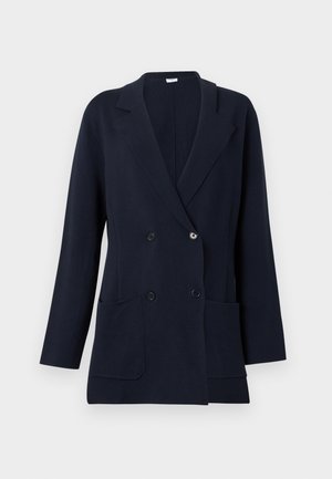 Frau trägt einen schwarzen zweireihigen Blazer, weißes Hemd, weite blaue Jeans, schwarze Absätze und hält eine gefaltete rote Clutch.