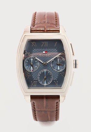 Ρολόι χειρός Tommy Hilfiger με ορθογώνια ασημένια κάσα, καφέ λουράκι από δέρμα, μπλε υφασμένο καντράν, ρωμαϊκούς αριθμούς και τρεις υποκαντράν για ημέρα, ημερομηνία και 24ωρη ένδειξη.