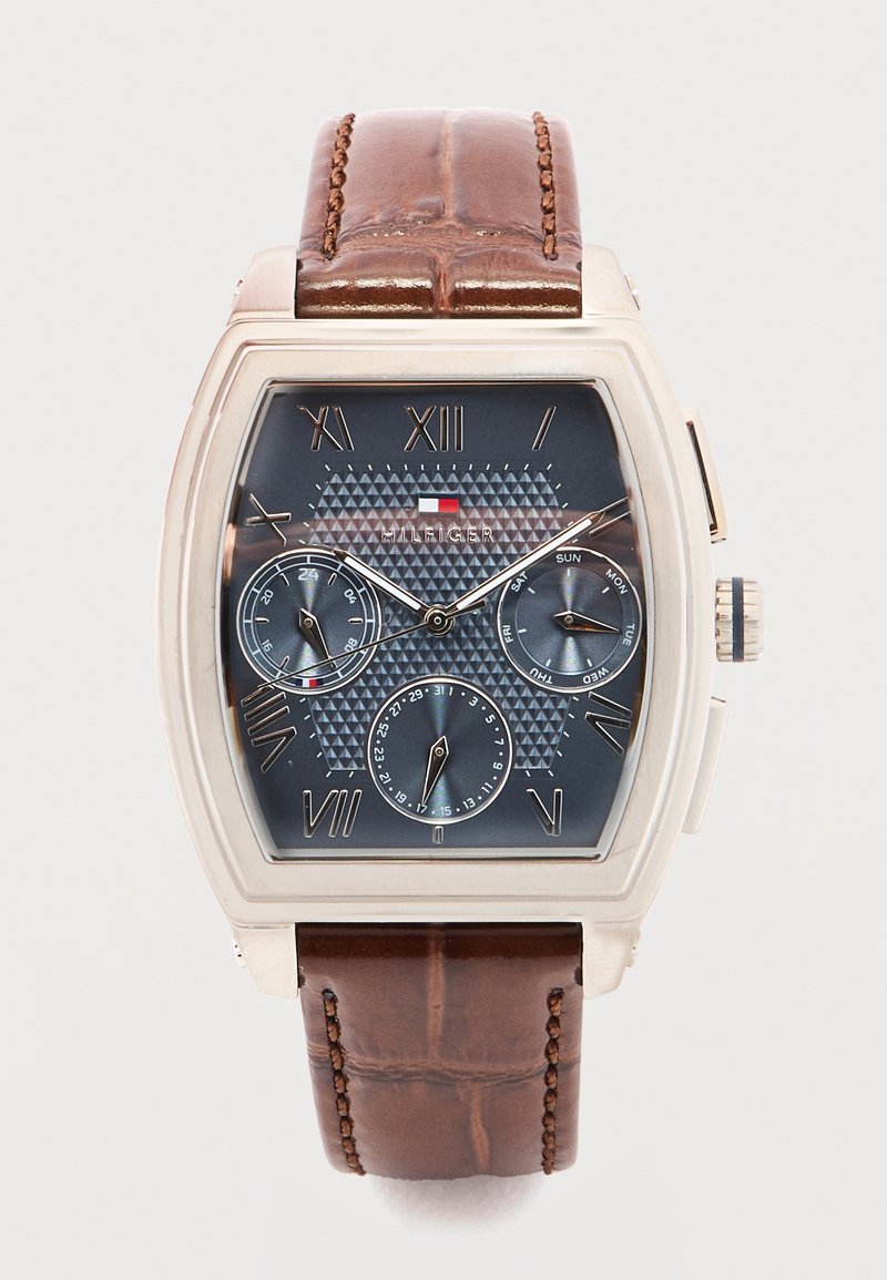 Ρολόι χειρός Tommy Hilfiger με ορθογώνια ασημένια κάσα, καφέ λουράκι από δέρμα, μπλε υφασμένο καντράν, ρωμαϊκούς αριθμούς και τρεις υποκαντράν για ημέρα, ημερομηνία και 24ωρη ένδειξη.
