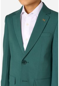 Blazer verde acqua con revers a punta, chiusura con un bottone e due tasche laterali. Presenta una camicia bianca sotto e un accento arancione sul colletto.