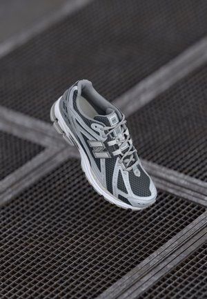 Chaussure de course New Balance grise et blanche suspendue au-dessus d'une surface métallique en treillis avec un motif hachuré en arrière-plan.