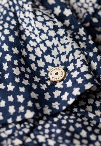 Tejido azul marino con un patrón de flores blancas en forma de estrella, que presenta una etiqueta circular dorada con el texto "SUPERDRY LOVE FROM" unida por un hilo negro.