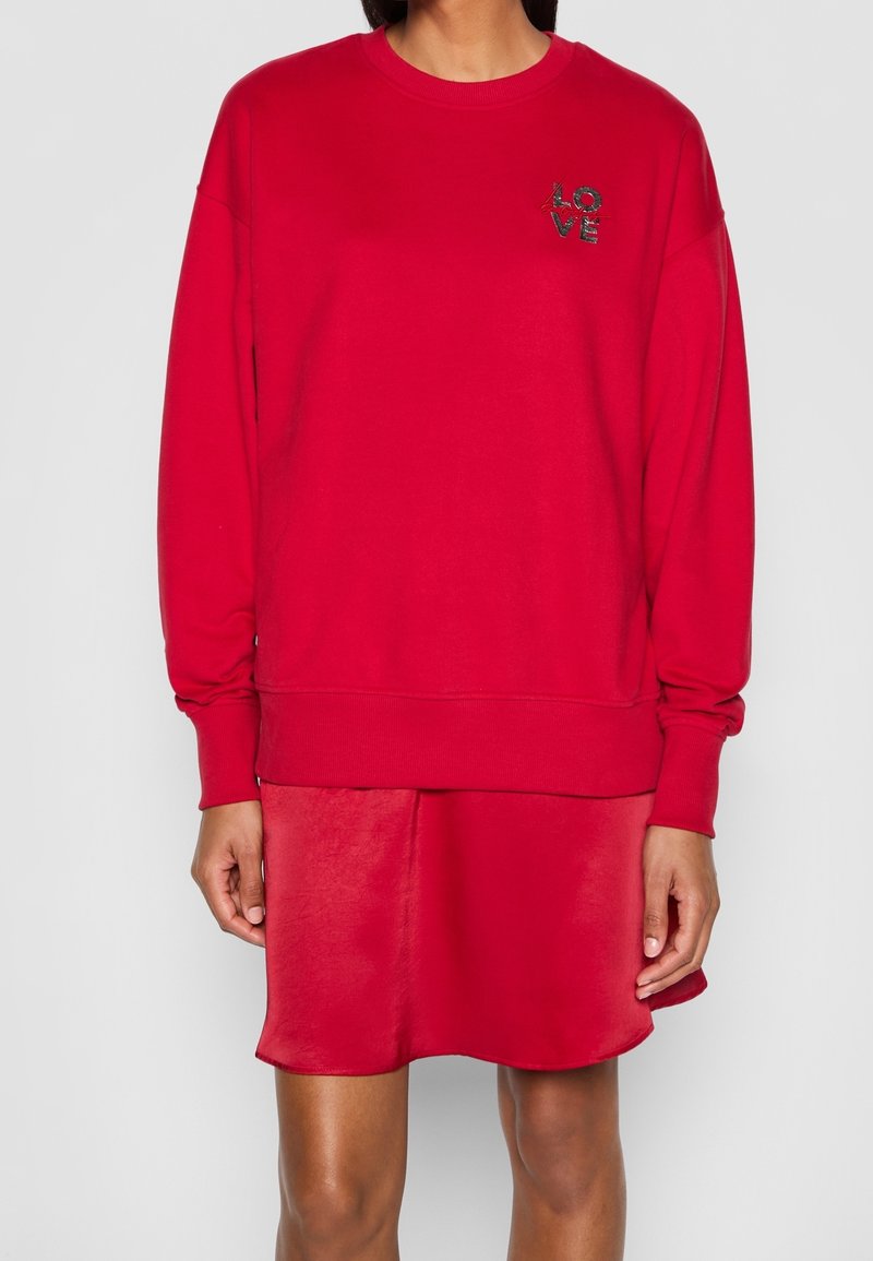 Sweat-shirt rouge en matière douce avec col rond, épaules tombantes et poignets côtelés. Dispose d'une broderie "LOVE" en couleurs contrastées. Porté sur une robe rouge assortie.