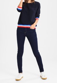 Marineblaues Sweatshirt mit rot-weißen und blauen gestreiften Akzenten an den Ärmelbündchen und dem Saum, kombiniert mit dunklen Skinny-Jeans und weißen Sneakers.