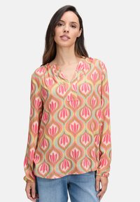 Femme portant un blouse à motifs rose, vert et beige à manches longues et un jean bleu clair, se tenant debout devant un fond blanc uni.