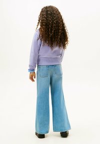 Fioletowy krótki sweter z ribbowymi mankietami, w połączeniu z szerokimi, jasnoniebieskimi dżinsami. Modelka ma kręcone włosy i jest odwrócona tyłem.