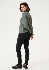 Green long-sleeve shirt with high collar, gathered shoulders, paired with black slim-fit jeans and glossy black shoes, standing against a plain background.