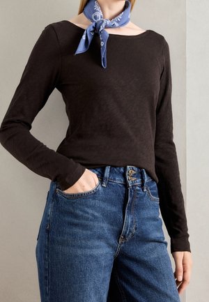 Personne portant une chemise à manches longues noire, un jean bleu à double fermeture par bouton, et un foulard bleu à motifs autour du cou, main dans la poche.