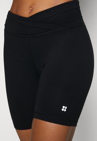 Black athletic shorts with a high, crossed waist design. Made of smooth fabric, featuring a small white logo on the lower side.