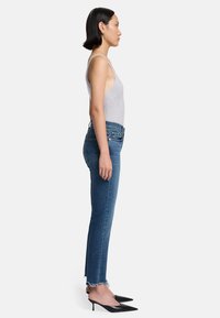 Canottiera grigia a coste, jeans skinny blu a vita alta con orli sfrangiati e décolleté neri a punta, visti di lato.
