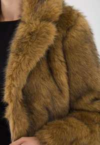Brun faux fur frakke med lange, bløde fibre, der har en bred krave og overlappende frontsektioner. Glat tekstur med subtil farvevariation.