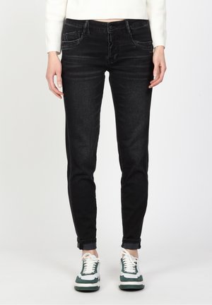 Personne portant un jean noir ajusté retroussé aux chevilles, des baskets blanches et vertes, et un haut blanc à manches longues court à la taille.