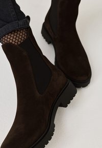 Botas Chelsea de ante marrón con paneles laterales elásticos, suelas de goma resistentes y una textura suave. Medias de red visibles por encima del tobillo.