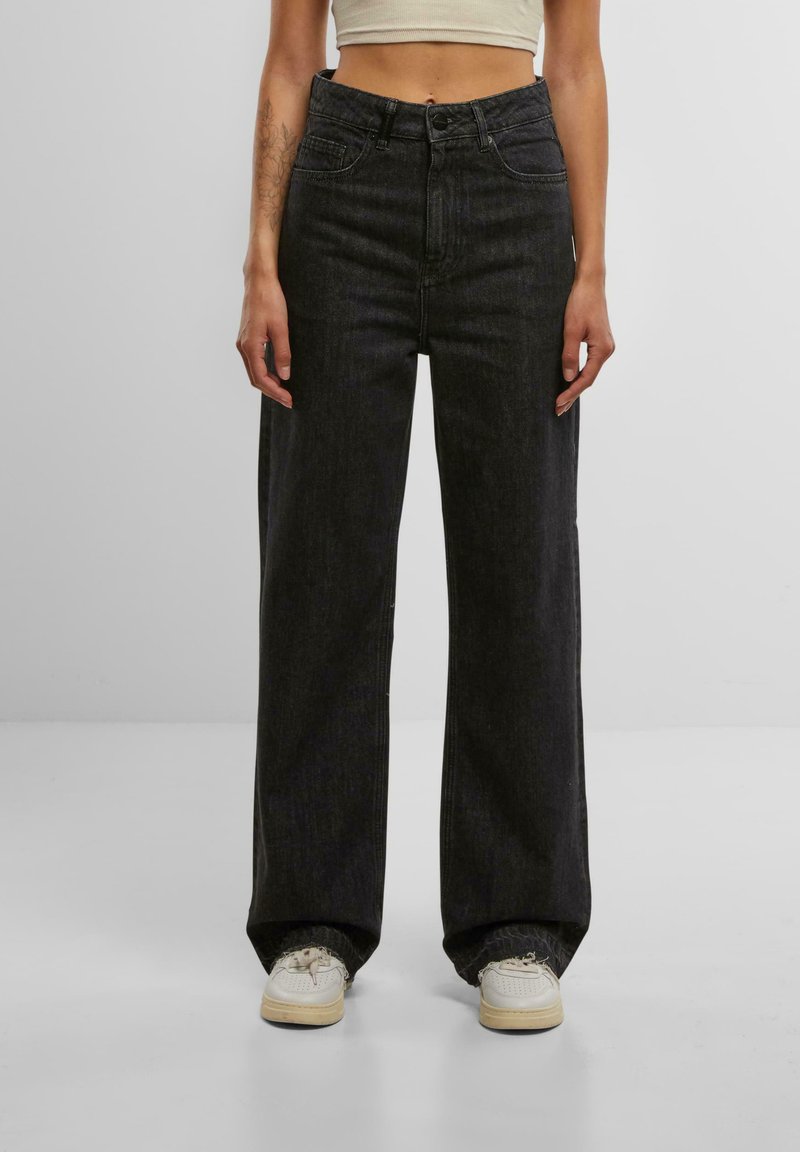 Schwarze Jeans mit weitem Bein und hoher Taille, ausgestattet mit Fronttaschen und einem Reißverschluss. Kombiniert mit weißen Sneakern, die einen lässigen Look unterstreichen.