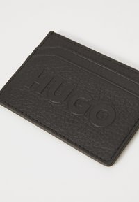 Cardholder din piele texturată neagră, cu logo-ul „HUGO” embosat, având trei sloturi pentru carduri și margini cusute pentru durabilitate.